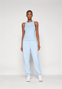 Mulher em pé de frente, vestindo uma parte de cima de tanque Adidas azul clara, calças de jogging combinando e ténis brancos, contra um fundo simples.