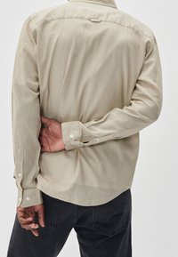 Beige long-sleeve shirt with a relaxed fit, textured fabric, and buttoned cuffs. Features curved hem and a back yoke detail.