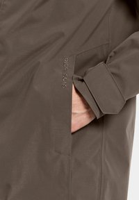 Brun vandtæt jakke med en glat tekstur, der har en sidelomme og broderet "Didriksons" logo på forsiden.