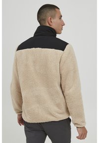 Beige fleece pullover with black collar, textured fur-like material, and loose fit. The back features a solid black panel across the shoulders.