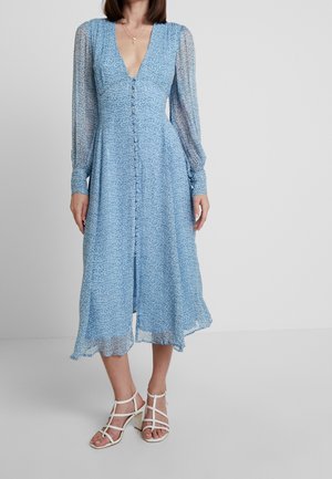 Sinivalkoinen kuvioitu mekko, jossa syvä V-pääntie, pitkät läpikuultavat hihat ja napituskiristys edessä. Liehuva helma, jota täydentävät valkoiset remmikorkokengät.