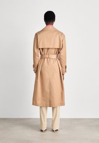 Beige trenchcoat met een riem in de taille, rugventilatie en kraagdetails, gecombineerd met crèmekleurige broek, gezien vanaf de achterkant.