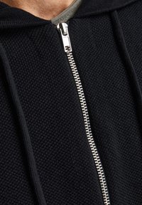 Black knitted hoodie with a silver zipper, featuring a textured surface and a well-defined hood. Underneath, a gray crew neck shirt is visible.