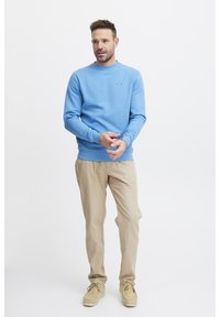 Sudadera azul claro con escote redondo, mangas largas y un pequeño detalle de logo. Pantalones beige y zapatos marrón completan el conjunto.