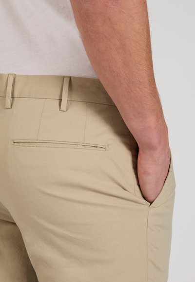 Main droite dans la poche d’un pantalon beige, poche arrière visible et passants de ceinture, portant une chemise blanche sur un fond uni clair.