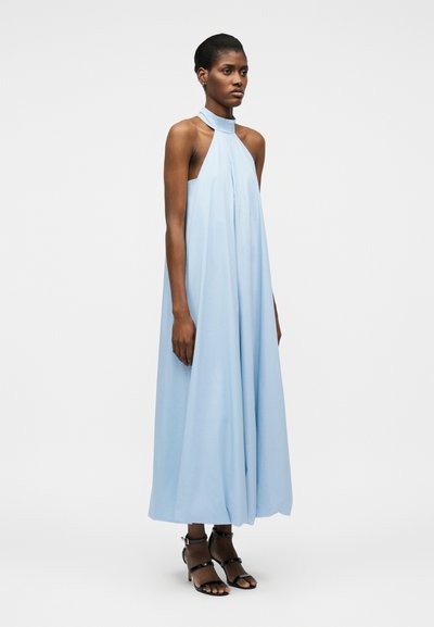 Femme mince portant une robe maxi bleu clair sans manches, ample, avec un col montant, et des sandales à talons noires à lanières, debout devant un fond blanc.