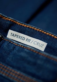 Tummansininen farkkukangas, jossa on kapeampi istuvuus. Huomiota herättävä oranssi ommellus reunoilla. Siisti, teksturoitu pinta.