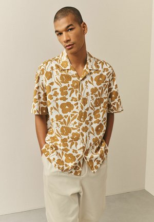 Mann mit kurzen Haaren, der ein beige-braunes kurzes Hemd mit Blumenmuster und hellfarbene Hosen trägt, mit in den Taschen stehenden Händen.