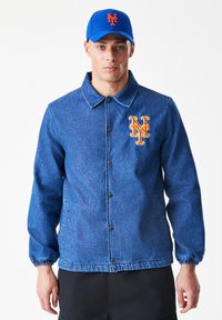 Džínová bunda v modré barvě, s límcem, patentními knoflíky a žebrovanými manžetami. Na hrudi je vyšitý týmový logo v oranžové barvě.
