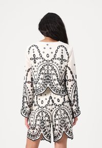 White embroidered outfit featuring intricate black floral designs, long sleeves, and a scalloped hem, with a loose fit and textured fabric.