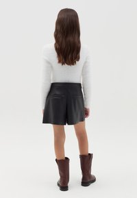 Pantaloni corti di pelle nera con tasche posteriori, abbinati a stivali marroni fino al ginocchio e a un maglione bianco peloso, visti da dietro.