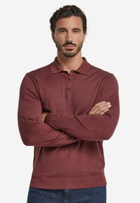 Mörkröd långärmad polo skjorta med krage, treknapps placket, ribbade ärmslut och en mjuk, texturerad yta. Bärs med armarna korsade.