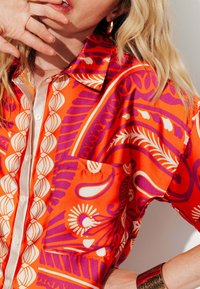 Chemise orange vif avec des motifs floraux blancs et violets, dotée d'un col pointu, d'une fermeture boutonnée et d'une poche sur le côté gauche.