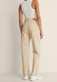 Pantalones de algodón beige con un diseño de pierna recta, bolsillos traseros visibles y una etiqueta de marca marrón en la cintura. Llevados con una camiseta sin mangas blanca.