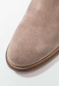 Botte en daim beige clair à texture lisse, bout rond et coutures visibles le long de la semelle. Présente un bord légèrement relevé de couleur marron.