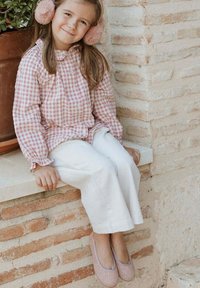 Niño con una camisa de cuadros rosa y pantalones blancos, con orejeras de peluche rosa y zapatos rosa claro adornados con puntos blancos.