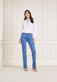Camicia bianca con maniche arrotolate, abbinata a jeans blu a gamba dritta e sandali beige con tacco, su uno sfondo chiaro e texturizzato.