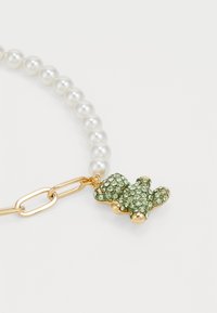 Colar de pérolas com uma corrente em tom dourado, apresentando um charme de urso de pelúcia decorado com cristais verdes, destacando uma superfície texturizada e acabamentos brilhantes.