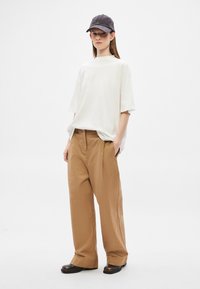 Za duży biały bawełniany t-shirt w połączeniu z luźnymi brązowymi spodniami, noszący ciemną czapkę i ciemne buty. Minimalistyczny design i luźny fason.