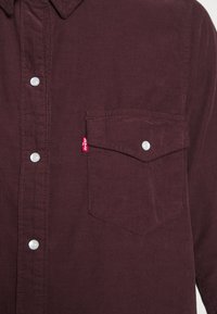 Chemise en velours côtelé bordeaux avec col pointu, deux poches poitrine et boutons-pression en plastique ; comporte un petit onglet Levi's rouge sur la poche gauche.