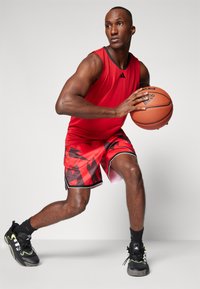 Jersey de baloncesto rojo con acentos negros, pantalones cortos a juego de rayas rojas y negras, y zapatillas deportivas negras con detalles verdes. Sosteniendo un balón de baloncesto.