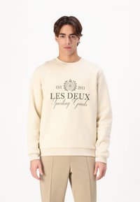 Crème-kleurige sweatshirt gemaakt van zacht materiaal, met een ronde halslijn en ribbelmanchetten. Cenraal geplaatste grafische tekst in donkergroen met de tekst "LES DEUX Sporting Goods."