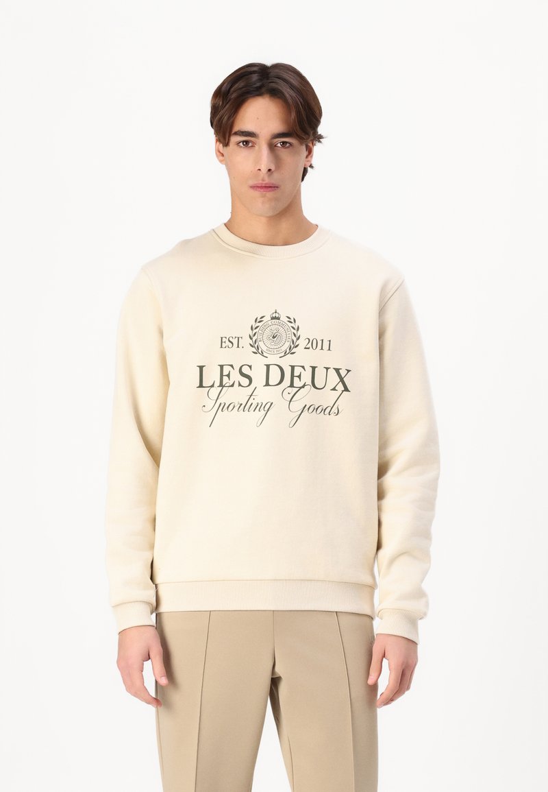 Crème-kleurige sweatshirt gemaakt van zacht materiaal, met een ronde halslijn en ribbelmanchetten. Cenraal geplaatste grafische tekst in donkergroen met de tekst "LES DEUX Sporting Goods."