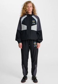 Svart atletisk träningsdräkt med grå accenter. Jackan med dragkedja har logotyp och elastiska muddar; byxorna har avsmalnande ben och logotypdetalj. Lättviktigt material.