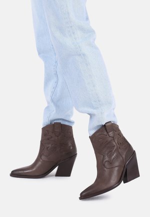 Osoba ubrana w jasnoniebieskie dżinsy i ciemnobrązowe skórzane botki na słupku z ozdobnym przeszyciem.