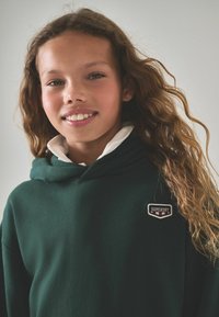 Felpa con cappuccio verde scuro realizzata in tessuto morbido con una patch logo ben visibile. Il colletto è bianco, aggiungendo un contrasto al design.