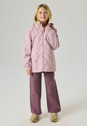 Laps, kellel on blondid juuksed, seljas heleroosa kapuutsiga jakk südamekujundustega, lilla laia jalaga püksid ja beežid tossud, seisab neutraalsel taustal.