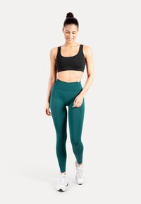 Zwarte sportbeha en teal hoge taille legging met een zijzak, gemaakt van rekbare stof, gecombineerd met witte sportschoenen.