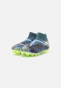 Soccer cleats with a dark upper featuring blue and green geometric patterns, knit collar, and a bright green textured sole with studs.