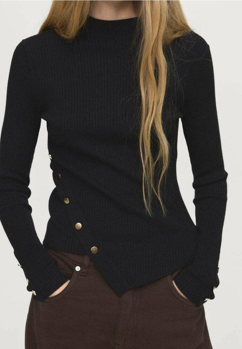 Femme portant un pull noir côtelé avec des boutons dorés sur le côté et un pantalon marron, mains dans les poches, longs cheveux blonds sur l'épaule.