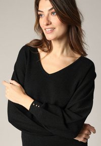Pull en tricot noir avec un col en V, un tissu texturé et des manches longues dotées de boutons en or aux poignets. Design lisse et ajusté.