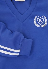 Blauwe katoenen V-hals sweater met wit geborduurd logo en lauwerkransontwerp. De manchetten zijn gedetailleerd met witte strepen voor textuur en accent.