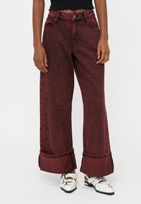 Pantaloni in denim rossi a gamba larga e vita alta, con una trama sbiadita, orli arrotolati e dettagli di cucitura in vista, abbinati a scarpe bianche slip-on.
