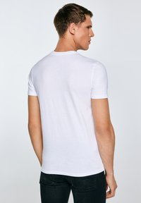Man die van achteren te zien is, draagt een effen wit T-shirt met korte mouwen en zwarte jeans, tegen een lichtgrijze achtergrond.