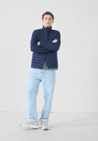Joven de pie con los brazos cruzados, vistiendo una chaqueta acolchada azul marino, camiseta gris, jeans claros y zapatillas blancas, frente a una pared lisa.
