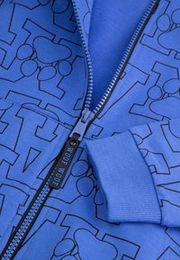 Niebieska bluza z kapturem na zamek błyskawiczny z grafiką czarnych odcisków łap oraz teksturowanym ściągaczem. Zamek z etykietą "WOOF WOOF". Miękki, elastyczny materiał.