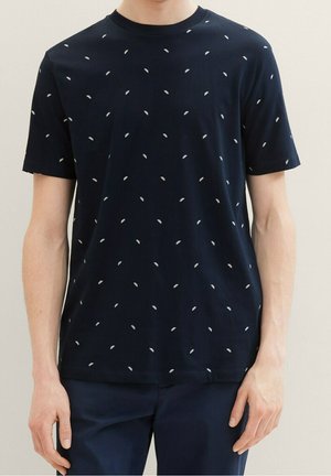 Mand iført en mørkeblå t-shirt med små hvide trykte former og matchende mørkeblå bukser, stående mod en ensfarvet baggrund.