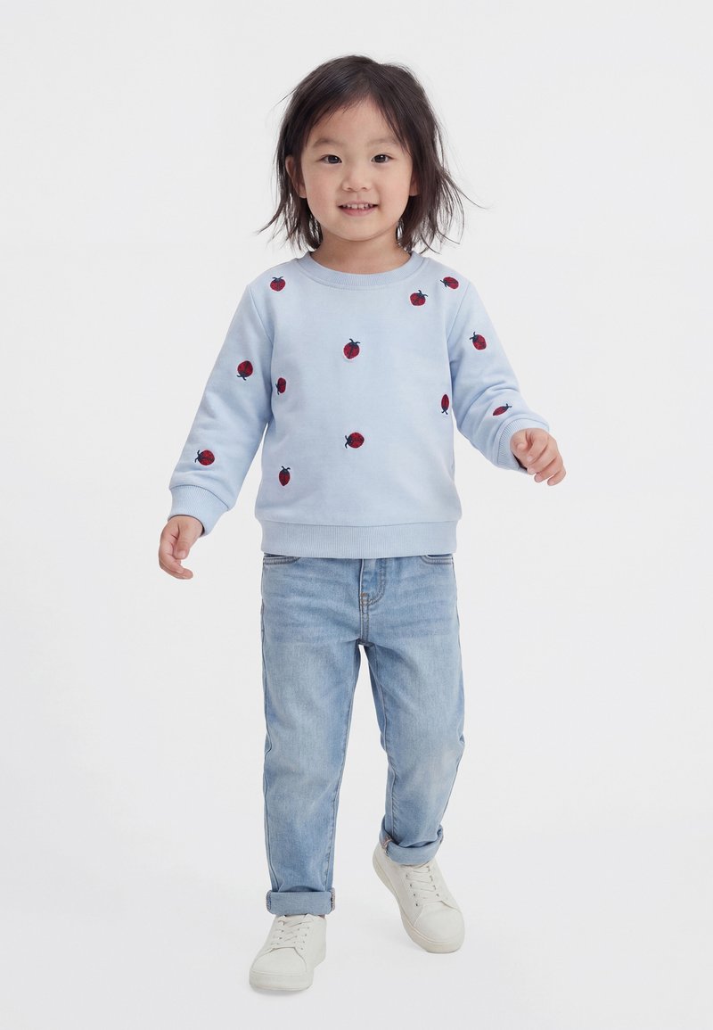 Enfant portant un sweat-shirt bleu clair avec des imprimés de coccinelles rouges, un jean bleu clair et des baskets blanches, debout devant un fond blanc uni.