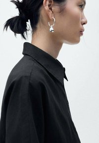 Silver hoop earring with a teardrop pendant, worn by a person in a black collared shirt, featuring soft fabric and a structured collar.
