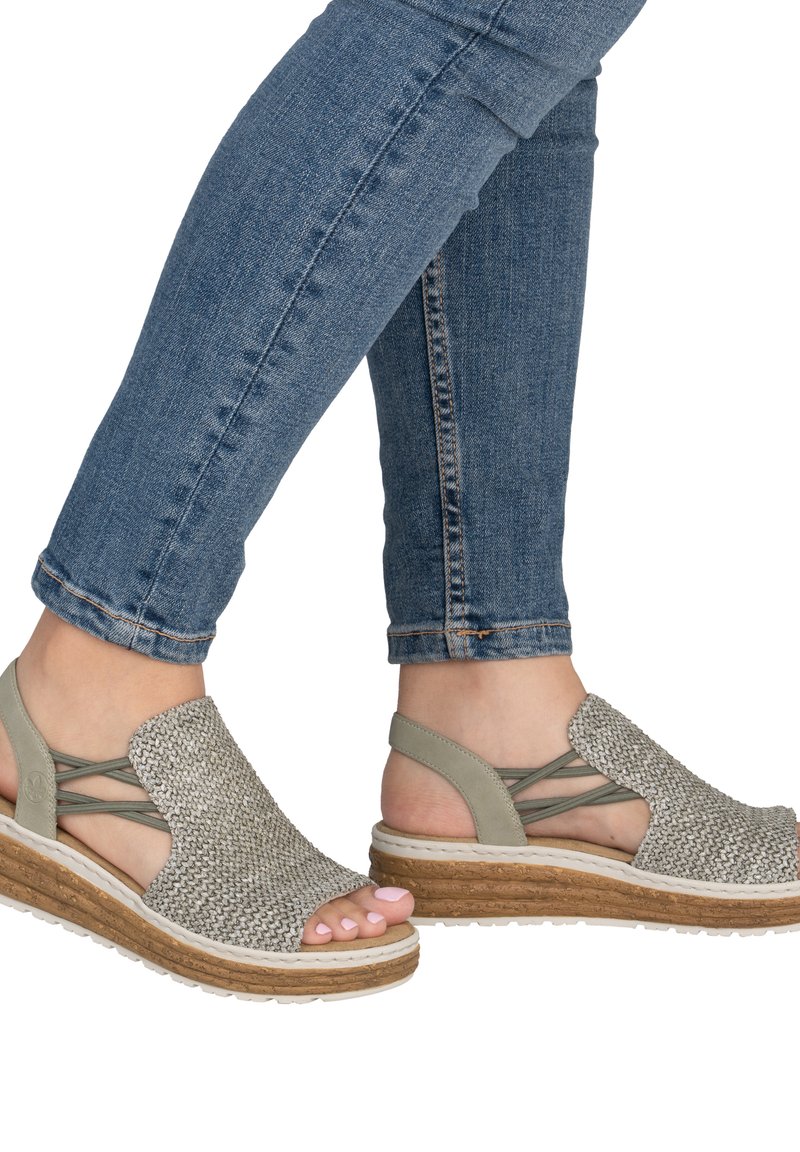 Pieds chaussés de sandales compensées en tissu gris avec des brides croisées, associées à un jean bleu roulotté.