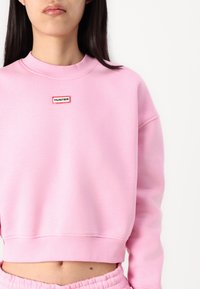 Pink cropped sweatshirt made of soft fabric, featuring a circular neckline and a small red and black "HUNTER" logo on the chest.