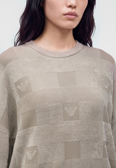 Femme aux cheveux foncés portant un pull beige texturé avec des motifs carrés subtils et des dessins en forme de V sur le tissu.