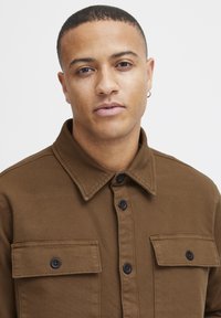 Camisa marrón de botones con cuello, dos bolsillos en el pecho y botones negros, hecha de un tejido texturizado. El modelo tiene el pelo corto y lleva un pendiente.