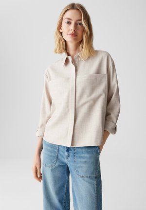Lichtbeige overhemd met knopen, grote borstzak, kraag en opgerolde mouwen, gecombineerd met lichtblauwe jeans.