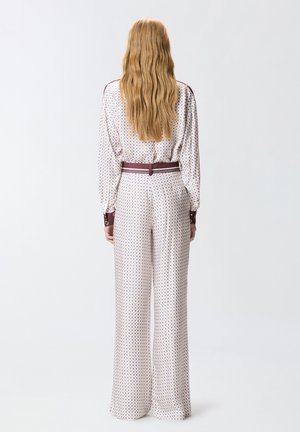 Kvinde med langt, bølget hår står med ryggen til iført hvide bukser med vide ben og en matchende langærmet bluse med små blå mønstre.