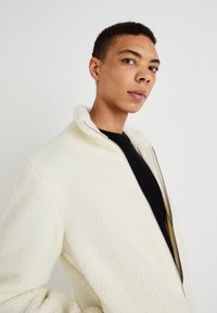 Casaco de fleece cor creme com um colarinho alto, fecho de correr à frente e tecido texturizado. Usado sobre uma blusa de malha preta.